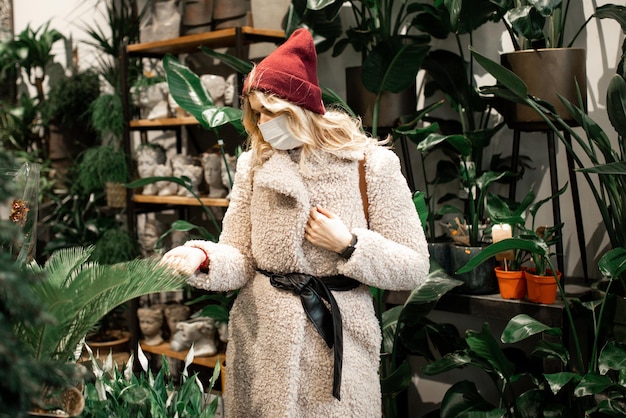 Young woman in faux fur coat and hat, wearing medical mask, chooses flowers in flower shop. New normal of social life, shopping. Gardening center, city life, lifestyle