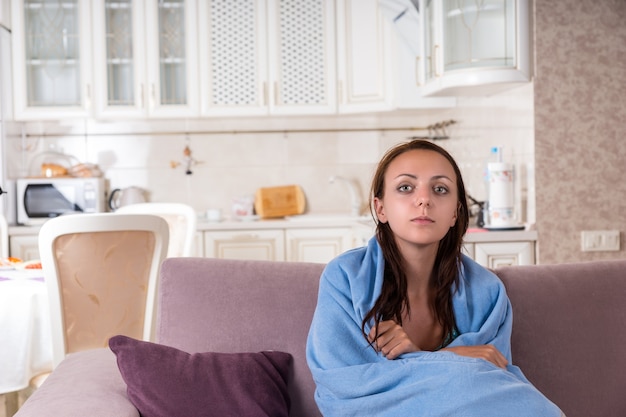 Giovane donna che si sente male o triste avvolta in un'accogliente coperta blu e seduta sul divano di casa, con lo sguardo fisso davanti a sé