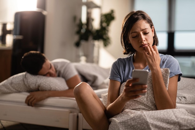 Young woman feeling uncertain while using mobile phone and reading text message Her boyfriend is sleeping on the bed behind her