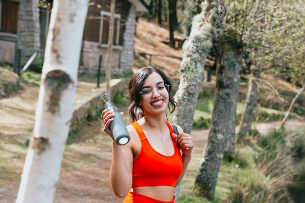 ヨガマットと水のボトルと公園でカメラに微笑んでフィットネス服を着た若い女性