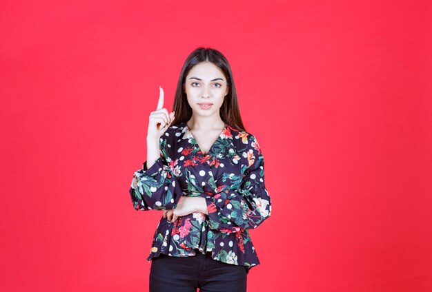 빨간 벽에 서서 거꾸로 보여주는 꽃무늬 셔츠를 입은 젊은 여자