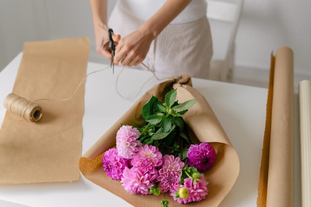 若い女性の花屋が、茶色のクラフト紙にダリアのピンクの花束を集め、デザイン用の止血帯の画像で結びます