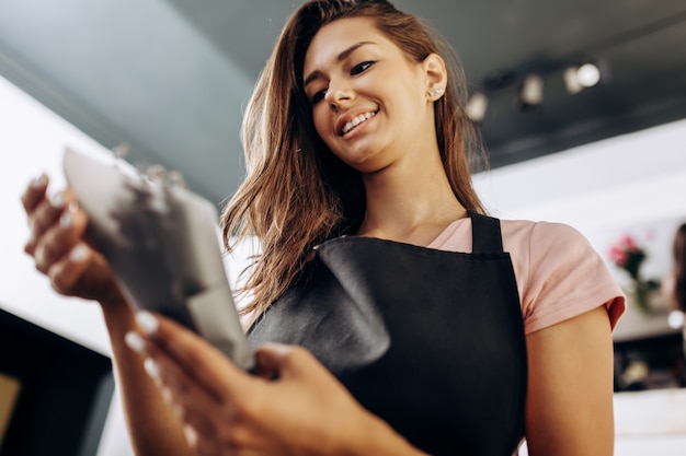 Young woman florist in a pink t-shirt and black apron holds a little bouquet of flowers packed in dark paper .