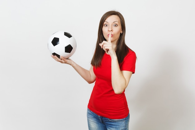 Young woman, football fan or player in red uniform hold soccer\
ball, say hush be quiet with finger on lips gesture isolated on\
white background. sport, play football, cheer, healthy lifestyle\
concept.