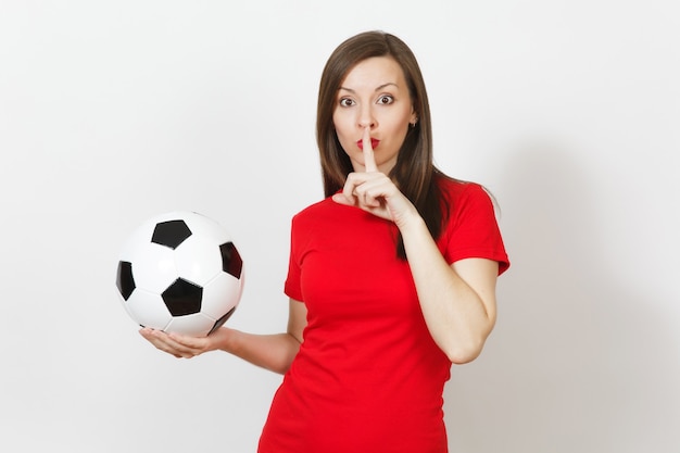 Young woman, football fan or player in red uniform hold soccer\
ball, say hush be quiet with finger on lips gesture isolated on\
white background. sport, play football, cheer, healthy lifestyle\
concept.