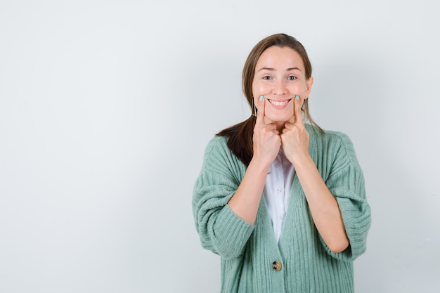 Young woman forcing a smile on face with fingers in blouse, cardigan and looking cheerful. front view.