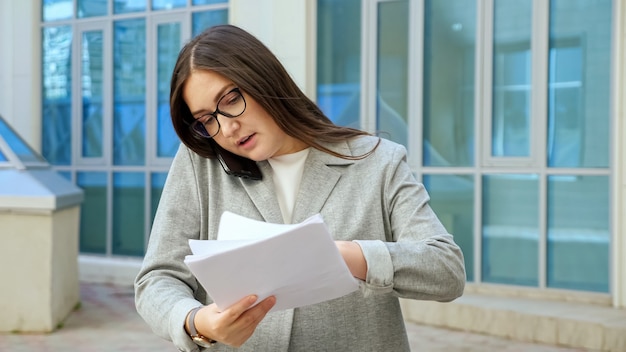 フォーマルなスーツと眼鏡をかけた若い女性が電話で話し、外出先で書類を調べます。