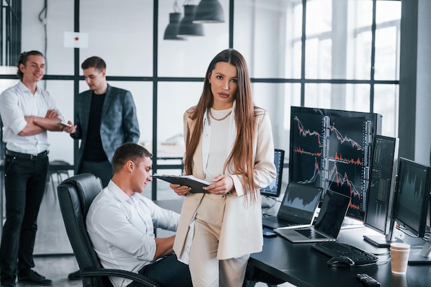 Young woman in formal wear standing of front of people Team of stockbrokers works in modern office with many display screens