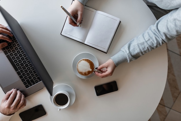 Photo young woman freelancer sits at a table with a business man in a cafe drinking coffee and making entries in a notebook