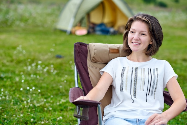 Young woman freelancer sitting on chair and relaxing in front of tent at camping site in forest or meadow.
