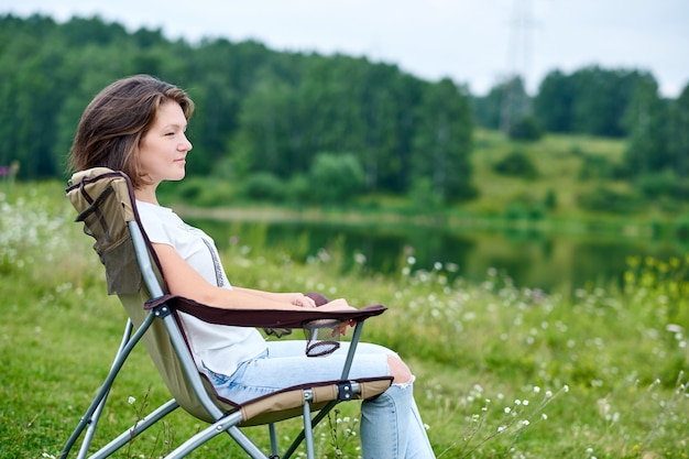 Young woman freelancer sitting on chair and relaxing in nature near the lake. Outdoor activity in summer. Adventure traveling in national park. leisure, vacation, relaxation