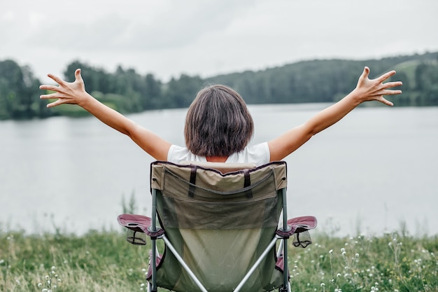 Young woman freelancer sitting on chair and relaxing in nature near the lake. Outdoor activity in summer. Adventure traveling in national park. leisure, vacation, relaxation