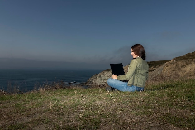 Foto giovane donna freelance seduta su una scogliera alta sopra il mare e che lavora a distanza