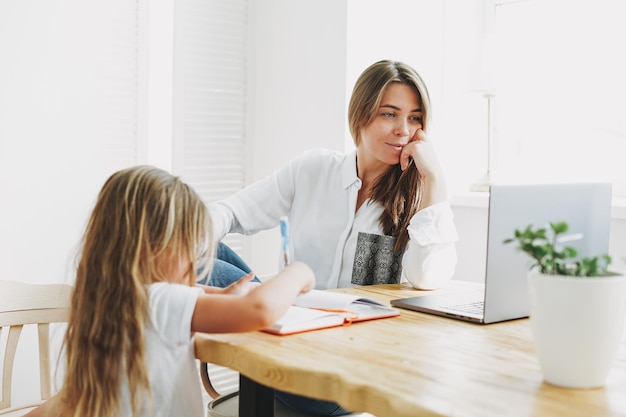 Young woman freelancer working on laptop with little daughter drawing on table at the home