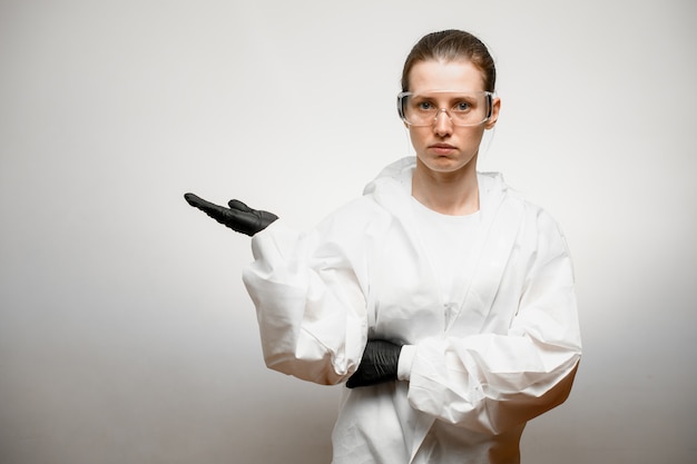 Young woman in full medical protective clothing stands on grey background and shows hand to side
