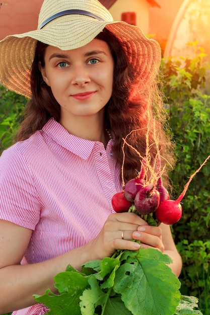 Young woman in the garden wearing hat and holding bunch of fresh radishes in sunny evening