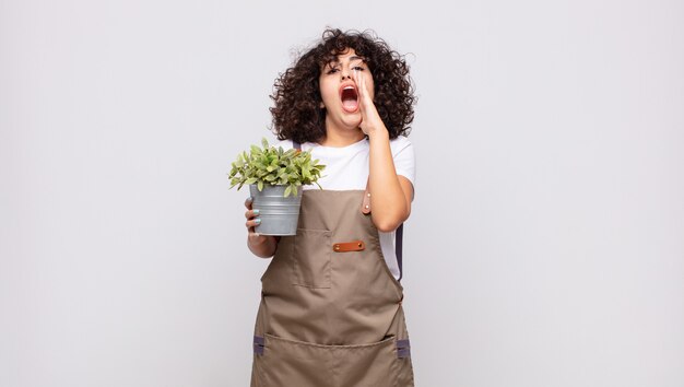 Young woman gardener feeling happy, excited and positive, giving a big shout out with hands next to mouth, calling out