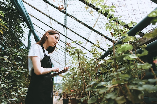 더 나은 품질 통제를 위해 정원 센터에서 일하는 디지털 태블릿을 가진 안경과 앞옷을 입은 젊은 여성 정원사