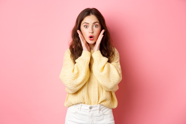 Young woman gasping and saying wow, looking surprised at camera, checking out promotion, staring at promo offer, standing against pink wall.