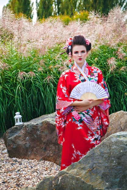 Giovane donna in costume da geisha con trucco di fantasia in giardino con un fan