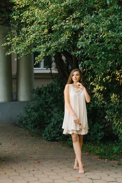 Young woman or girl in a flying light dress walks barefoot near a house with columns and green trees