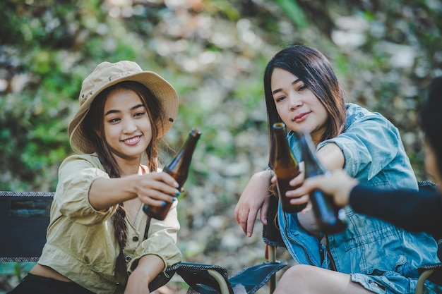 Young woman and girl friends travelers relaxing in camp chairs at tent They are cheering and drinking beer during camping talking with fun and happy together