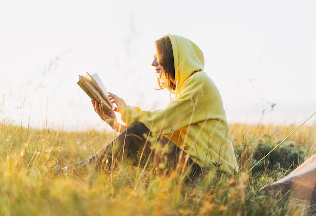 Young woman girl traveler in yellow hoodie reading book in morning sunrise