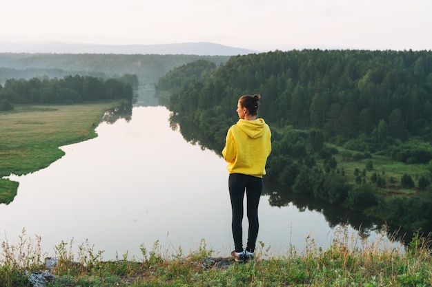 Young woman girl traveler in yellow hoodie with cup of morning coffee at sunrise on beautiful view