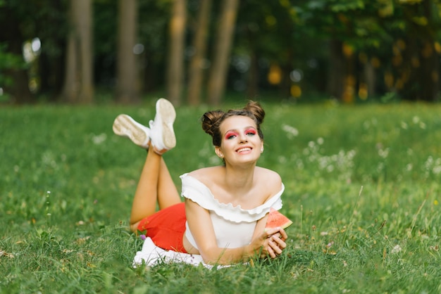 Молодая женщина или девушка с ярким макияжем держит в руках кусочек сочного арбуза и лежит на зеленой гра