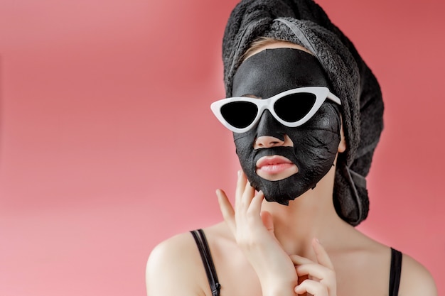 Young woman in glasses appling black cosmetic fabric facial mask. Face peeling mask with charcoal, spa beauty treatment, skincare, cosmetology. Close up