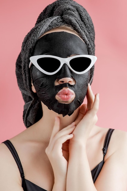 Young woman in glasses appling black cosmetic fabric facial mask on pink background. Face peeling mask with charcoal, spa beauty treatment, skincare, cosmetology. Close up.