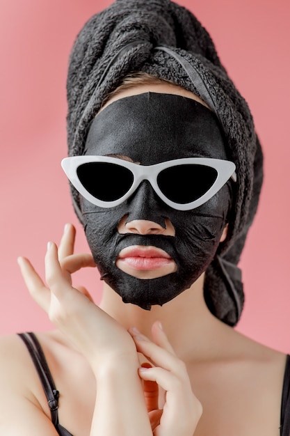 Foto la giovane donna in vetri applica la maschera facciale nera del tessuto cosmetico sulla parete rosa. maschera peeling viso con carbone, trattamenti di bellezza spa, cura della pelle, cosmetologia. avvicinamento