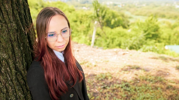 Young woman in glasses business suit standing near tree in nature Business woman posing near tree trunk during break on background of countryside