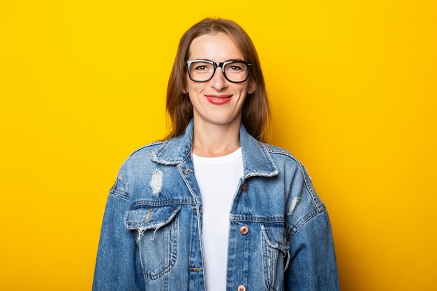 Young woman in glasses and denim jacket on yellow wall.