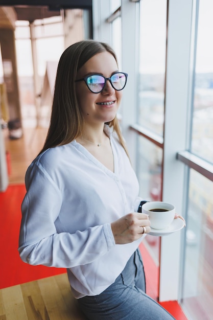 ヨーロッパ風のメガネをかけた若い女性は、白いシャツを着てコーヒーを飲み、窓の外を見ています。