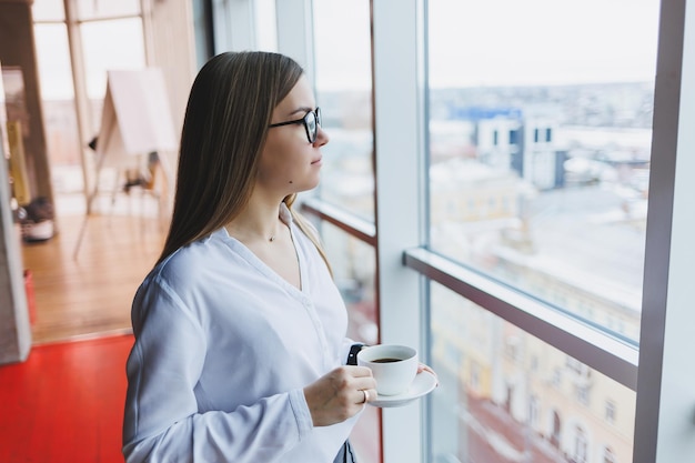 ヨーロッパ風のメガネをかけた若い女性は、白いシャツを着てコーヒーを飲み、窓の外を見ています。