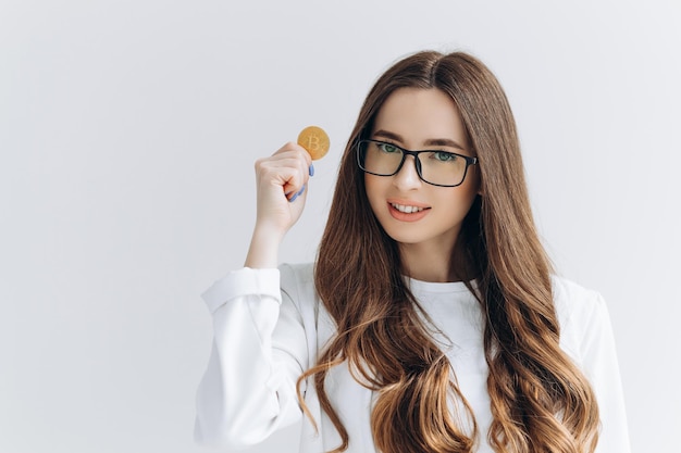 Young woman in glasses holding a Bitcoin isolated on grey background making money gesture