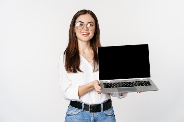 Young woman in glasses showing laptop screen, demonstrating promo on computer, website or store, standing over white background