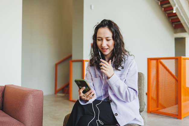 Young woman in glasses sitting in a chair in a modern cafe at a table with a laptop working online A woman is talking on the phone through headphones Remote work Work of a freelancer