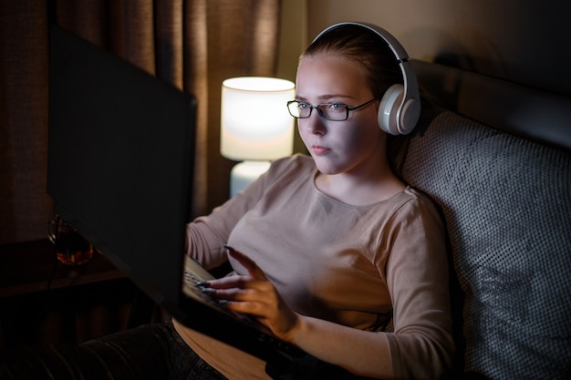 Young woman in glasses studying or working using laptop at night in home interior. Overtime sofa workspace. Attentive serious teenager student studies hard at night. Stress addiction to social media.
