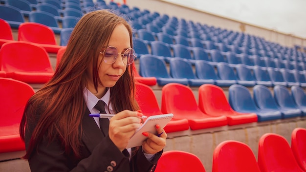 Young woman in glasses with notepad pen sitting on stadium bleachers alone Female journalist writing down notes during competitions at street stadium