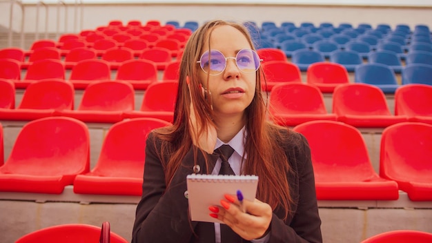 Young woman in glasses with notepad pen talking on mobile phone sitting on stadium bleachers Female journalist speaking on smartphone discussing competitions at street stadium