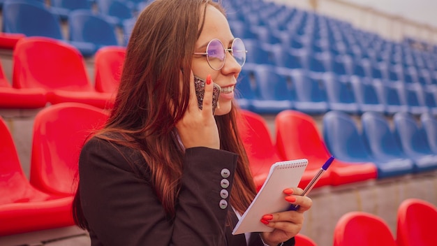 Young woman in glasses with notepad pen talking on mobile phone sitting on stadium bleachers