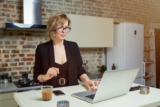 A young woman in glasses works remotely on a laptop in her kitchen. A serious girl calmly browsing news on the internet at home. .