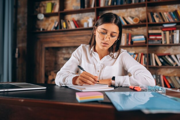 Photo young woman in glasses writes in a notebook while sitting at the table