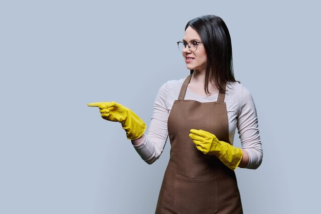 Young woman in glove apron pointing at empty copy space for text on grey background