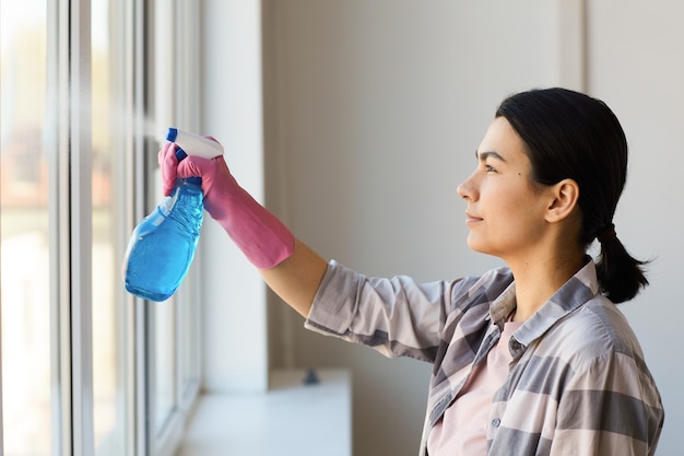 Young woman in gloves cleaning window with cleanser spray at home