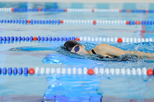 Giovane donna in occhiali e berretto da nuoto stile corsa strisciante anteriore nella piscina da gara coperta di acqua blu.