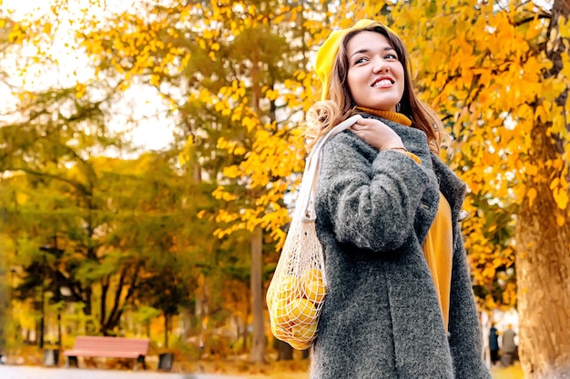 Молодая женщина в сером пальто в парке на открытом воздухе держит сумку ECOnet с фруктами и лимонами.