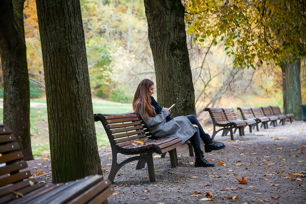 秋の都市公園のベンチに座って、彼女の携帯電話で検索している灰色のコートを着た若い女性